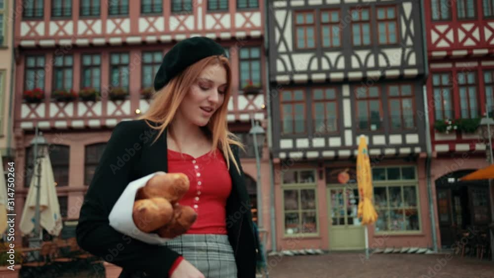 Beautiful, red-haired French girl in black beret holding paper-wrapped loaves in her hand smiles, go ahead, look around the left to the right. On street, outside Beautiful, red-haired French girl in black beret holding paper-wrapped loaves in her hand smiles, go ahead, look around the left to the right. On street, outside