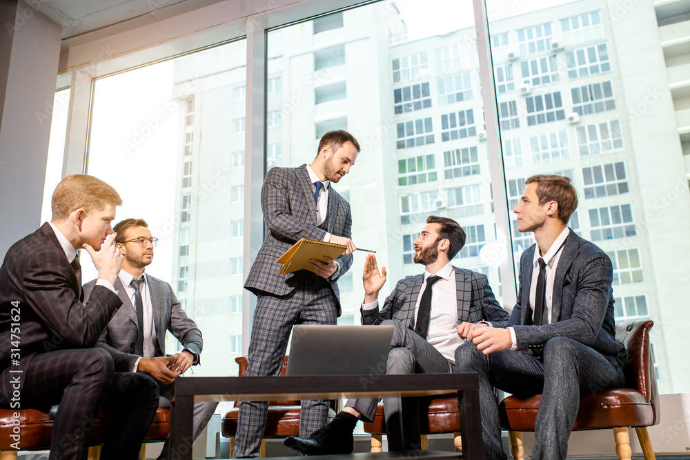 young caucasian business colleagues sit on leather chairs and discuss together in modern office, wearing elegant suits. big panoramic window in the background young caucasian business colleagues sit on leather chairs and discuss together in modern office, wearing elegant suits. big panoramic window in the background