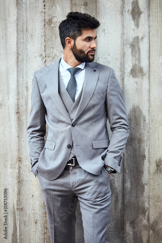 Adult caucasian brunet bearded businessman wear grey suit pose, keep hand in pockets, look away