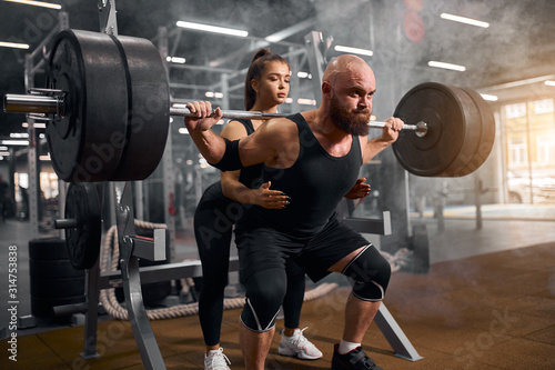 Supportive young pretty female trainer standing behind brutal hairless man, squatting with heavy barbell in brightly lighted fitness room, demonstrates right position, fitness performing