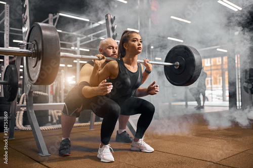 Cool attractive sporty fitness woman dressed in black sportswear, squatting using heavy barbell, taking help of professional fitness trainer, training in modern gym, preparing for competition