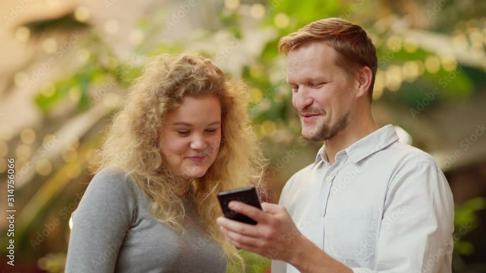 Medium waist up shot of happy couple looking at smartphone screen, having fun and laughing standing against blurred outdoor background Medium waist up shot of happy couple looking at smartphone screen, having fun and laughing standing against blurred outdoor background