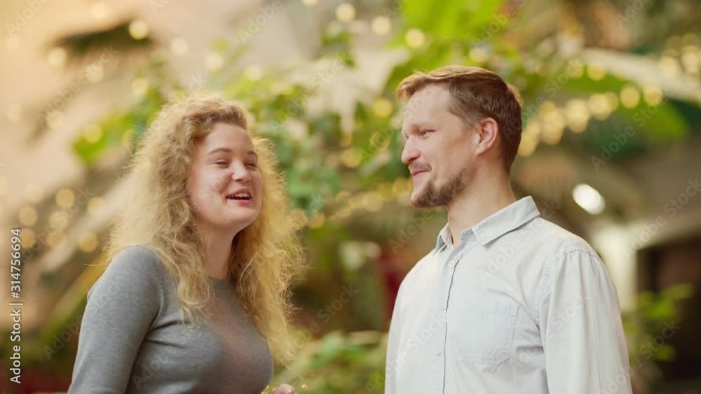 Waist up medium shot of positive middle aged man and cheerful young woman having joyful conversation and laughing while standing against blurred outdoor background with green plants and bright lights Waist up medium shot of positive middle aged man and cheerful young woman having joyful conversation and laughing while standing against blurred outdoor background with green plants and bright lights
