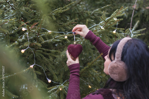Hands of brunette decorates a Christmas tree toy in the shape of a red heart  outdoor. The concept of decorating a Christmas tree, love, New Year and Christmas.