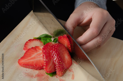 View of the hands of a girl holding a knife while preparing a fruit salad. Detail of the hands of a small girl while cutting strawberries with a kitchen knife.