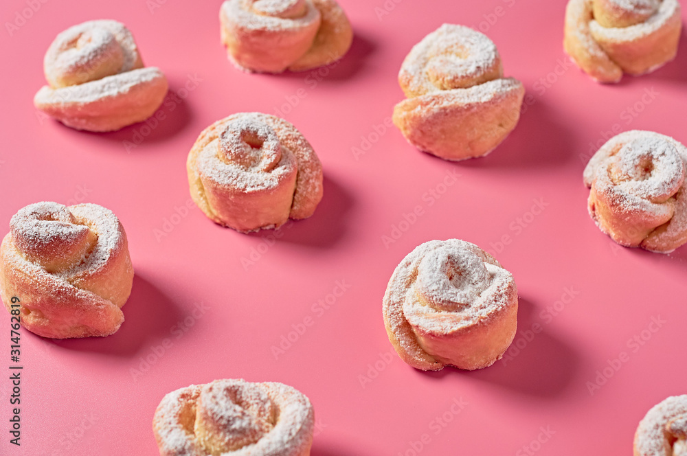 Rows of many homemade baked buns in form of rose spangled of powdered sugar lies on pink background. Close-up