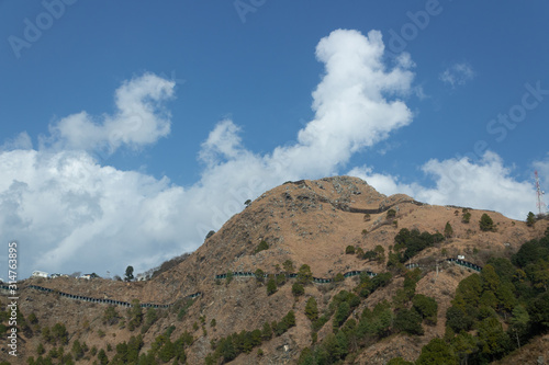 Low angle zoomed in wide shot of a mountain with shed covered roads and cloudy blue sky in the background.
