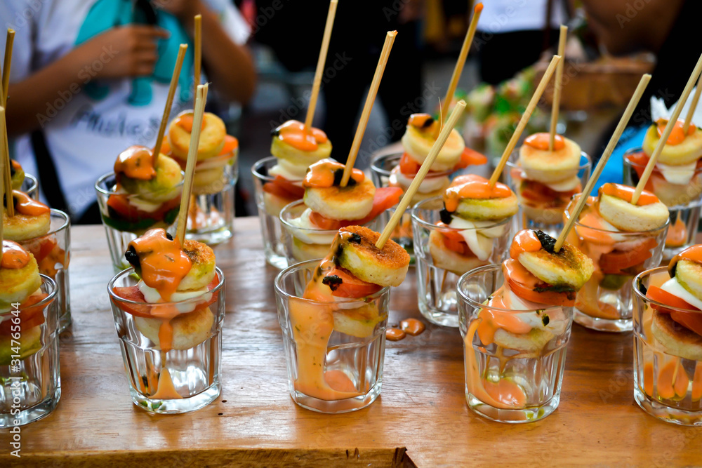 Snacks set on wooden table. People in the background tasting Street food. Seafood skewers with dressing in glass shots. Delicious luxury appetizer. Degustation, cooking festival, market. Close up. Snacks set on wooden table. People in the background tasting Street food. Seafood skewers with dressing in glass shots. Delicious luxury appetizer. Degustation, cooking festival, market. Close up.