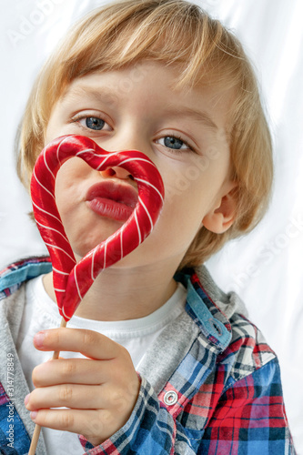 Cute little boy in with candy red lollipops in heart shape, white background. Beautiful kid eat sweets. Valentine's day, love concept. photo