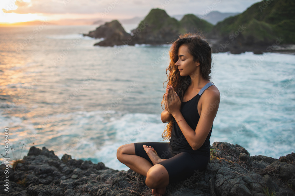 Young woman praying and meditating alone at sunset with beautiful ocean and mountain view. Self-analysis and soul-searching. Spiritual and emotional concept