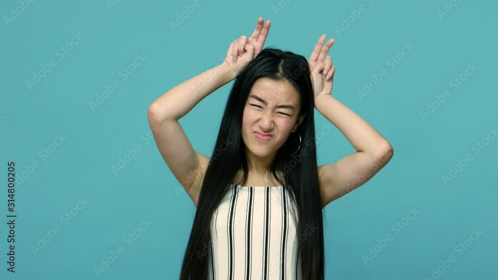 Angry asian woman with long hair threatening with bull horn gesture, looking at camera with hostile bully expression, provoking to conflict, attack. indoor studio shot isolated on blue background Angry asian woman with long hair threatening with bull horn gesture, looking at camera with hostile bully expression, provoking to conflict, attack. indoor studio shot isolated on blue background