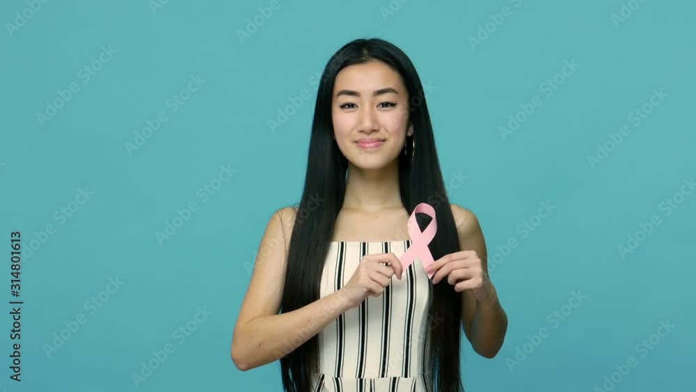Happy beautiful asian woman with long black hair holding pink ribbon, symbol of breast cancer awareness, female healthcare concept and medical insurance. indoor studio shot isolated on blue background Happy beautiful asian woman with long black hair holding pink ribbon, symbol of breast cancer awareness, female healthcare concept and medical insurance. indoor studio shot isolated on blue background