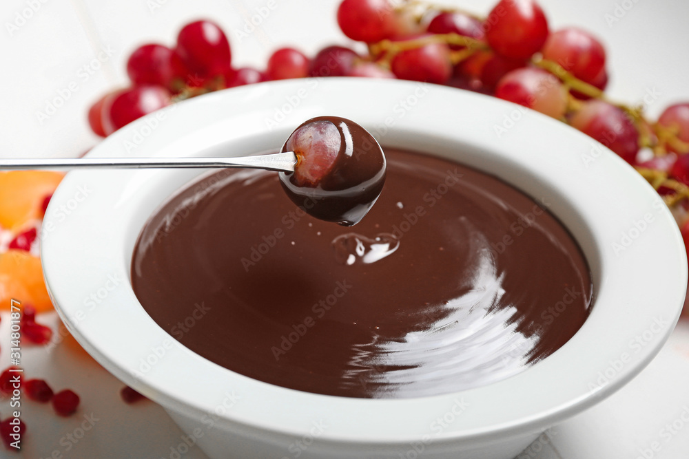 Dipping of tasty grape into bowl with chocolate fondue on table, closeup
