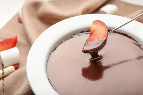 Dipping of tasty plum into bowl with chocolate fondue on table, closeup