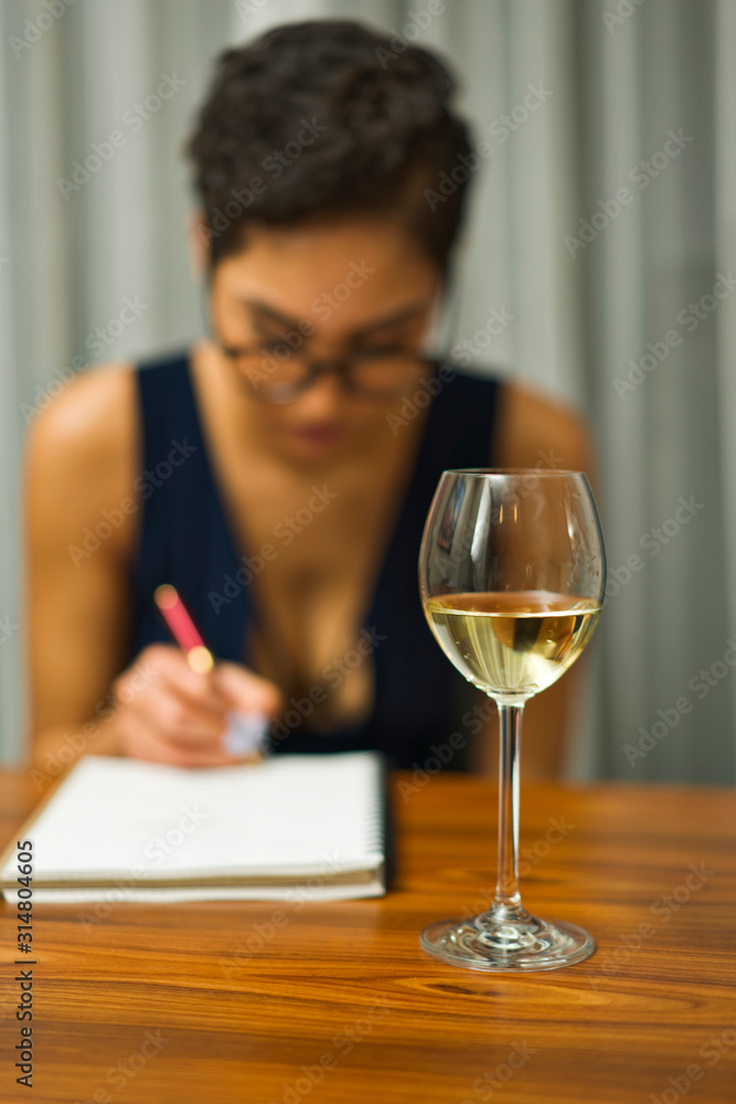A woman wearing glasses sits at a table drinking white wine and writing in a journal A woman wearing glasses sits at a table drinking white wine and writing in a journal