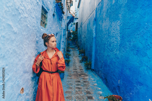 a tourist in a bright long African dress with a belted strap stands in a very narrow street of the blue city of Morocco © nelen.ru