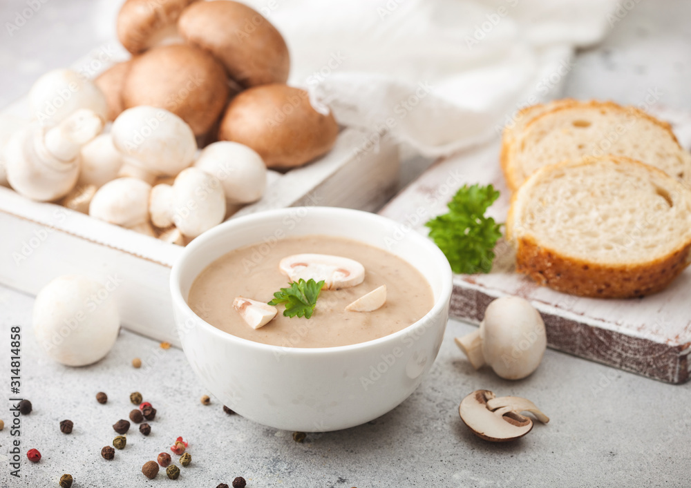 White bowl plate of creamy chestnut champignon mushroom soup on white kitchen background and box of raw mushrooms and fresh bread. White bowl plate of creamy chestnut champignon mushroom soup on white kitchen background and box of raw mushrooms and fresh bread.
