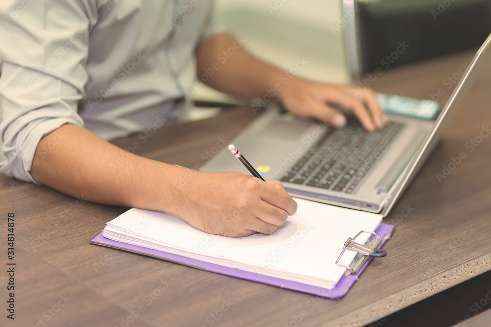 A man's hand is writing something on a white notebook and a laptop that looks to the side.Office workers writing text on white paper with mobile phones. A man's hand is writing something on a white notebook and a laptop that looks to the side.Office workers writing text on white paper with mobile phones.