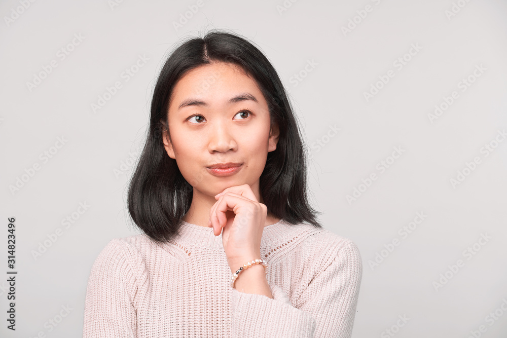 Young woman holding fingers to mouth chin. Thinks and analyzes important things about work study, looks up. Girl with brown eyes dressed in knitted cardigan stands isolated white background in Studio Young woman holding fingers to mouth chin. Thinks and analyzes important things about work study, looks up. Girl with brown eyes dressed in knitted cardigan stands isolated white background in Studio