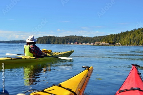 Group of friends on sea kayak in Pacific Ocean near Vancouver Island. Colorful kayaks, trees on the island, man with west, hat and paddle. Blue sky. 