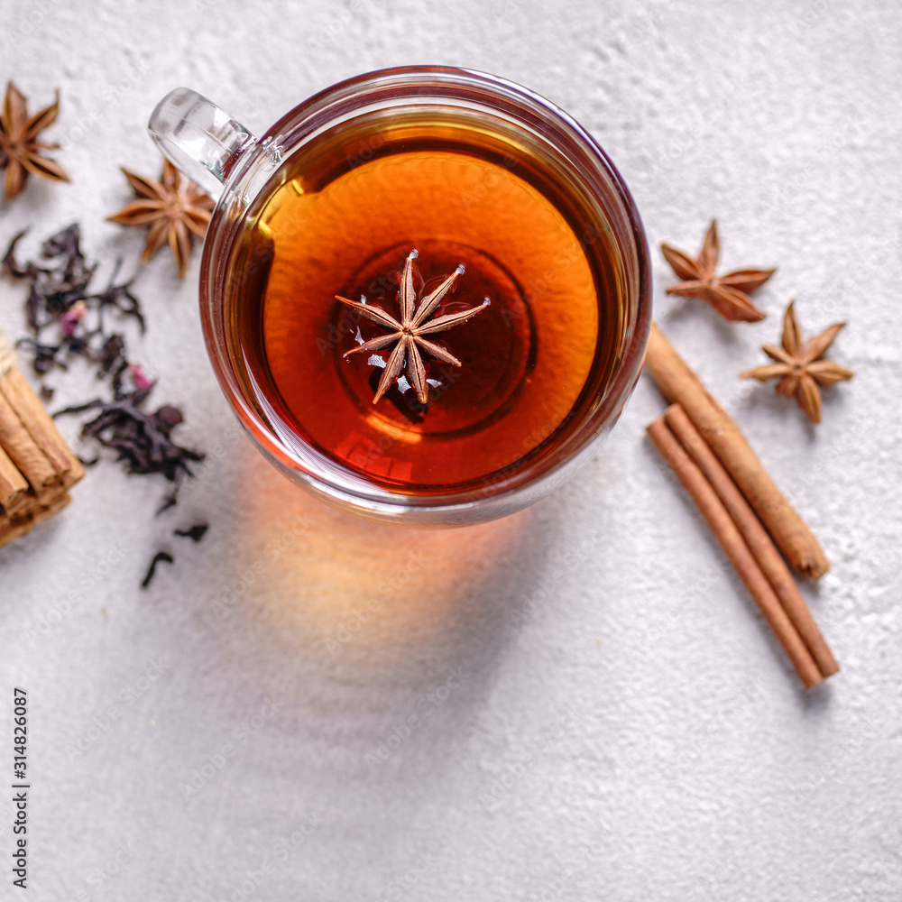 Fototapeta premium Spicy warming black tea in a glass cup with anise, cinnamon, tea stainer and sprinkled tea leaves on stone background. Cozy warming home atmosphere concept
