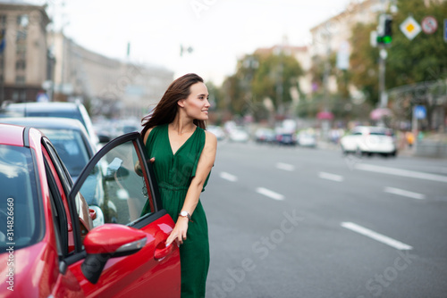 Fashion model wearing green overall posing outdoor next to her red car on the road. Young beautiful brunette caucasian woman driver. Beautiful girl, urban portrait. © Khorzhevska