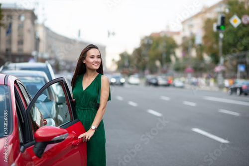 Fashion model wearing green overall posing outdoor next to her red car on the road. Young beautiful brunette caucasian woman driver. Beautiful girl, urban portrait. © Khorzhevska
