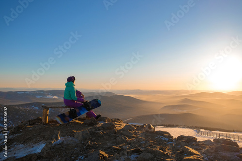 Girl snowboarder on top of a mountain in Sheregesh on the background of the sunset blue yellow sky. Hiking up the mountain.