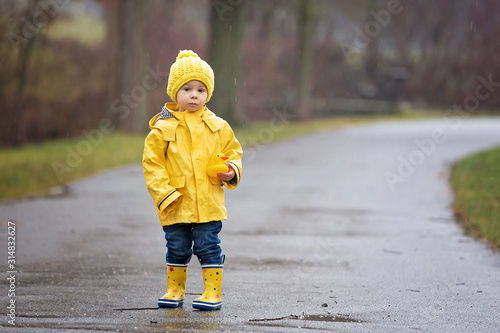 Beautiful funny blonde toddler boy with rubber ducks and colorful umbrella, jumping in puddles and playing in the rain