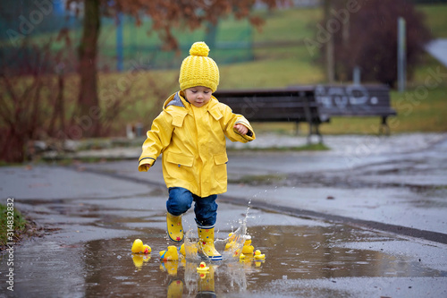 Fototapeta Naklejka Na Ścianę i Meble -  Beautiful funny blonde toddler boy with rubber ducks and colorful umbrella, jumping in puddles and playing in the rain