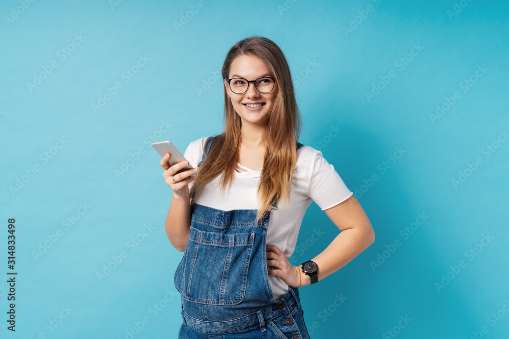 Smiling pregnant woman, with glasses, holding a mobile phone in one hand over blue background. Smiling pregnant woman, with glasses, holding a mobile phone in one hand over blue background.
