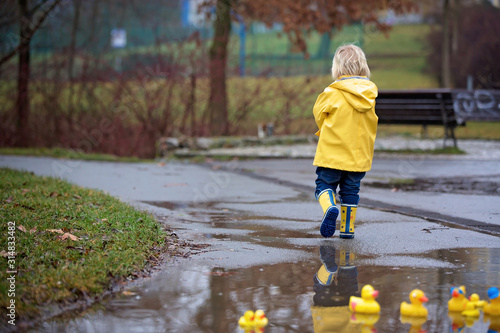 Beautiful funny blonde toddler boy with rubber ducks and colorful umbrella, jumping in puddles and playing in the rain