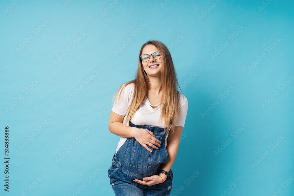Smiling pregnant woman wearing glasses caressing her belly over blue background Smiling pregnant woman wearing glasses caressing her belly over blue background