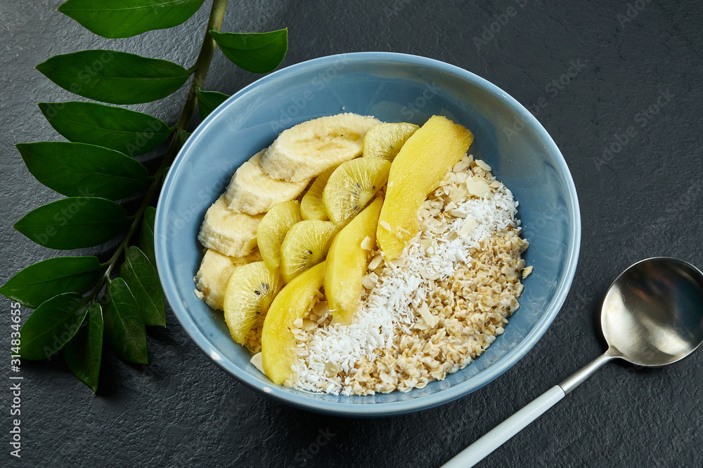 Close up view on Healthy breakfast: oatmeal with coconut, mango, bananas and yellow kiwi in a blue bowl on a black background. Close up view on Healthy breakfast: oatmeal with coconut, mango, bananas and yellow kiwi in a blue bowl on a black background.