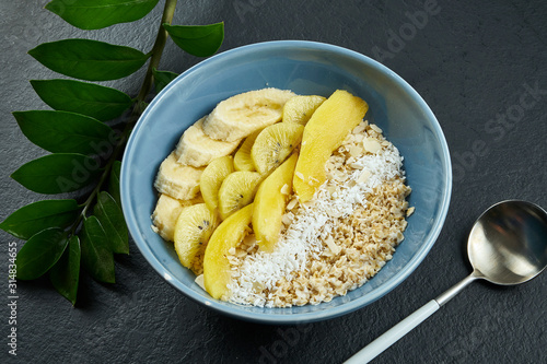 Close up view on Healthy breakfast: oatmeal with coconut, mango, bananas and yellow kiwi in a blue bowl on a black background.