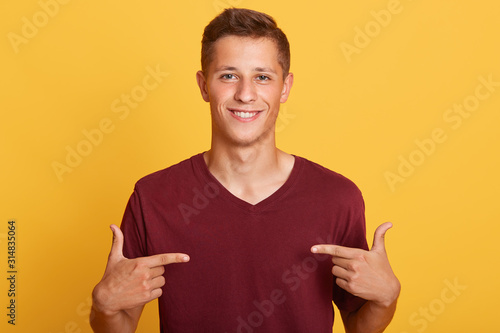 Close up of handsome man in blank burgundy t hirt pointing at himself isolated over yellow studio background, male with pleasant appearance looking smiling ditectly at camera. Advertising concept. photo