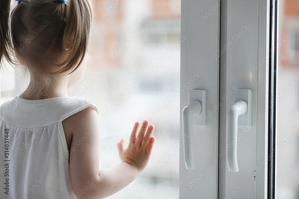 A little girl is sitting on the windowsill. A bouquet of flowers in a vase by the window and a girl sniffing flowers. A little princess in a white dress with a bouquet of white flowers by the window.