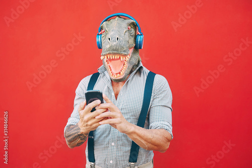 Senior fashion man wearing t-rex mask using mobile smartphone listening music with headphones - Crazy bizarre hipster guy having fun with new technology -  Funny and absurd surreal concepts photo