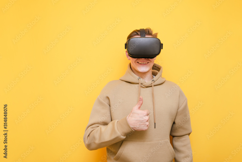 Happy young man in casual wear and VR helmet stands on a yellow background, looks into the camera with a smile and shows his thumb up, the guy likes virtual reality. Isolated. Copy space Happy young man in casual wear and VR helmet stands on a yellow background, looks into the camera with a smile and shows his thumb up, the guy likes virtual reality. Isolated. Copy space