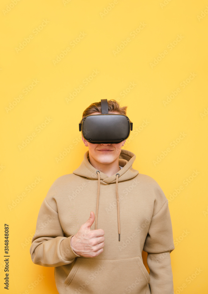 Positive teen guy in virtual reality helmet and casual clothes isolated on yellow background, showing thumbs up and smiling.Young man wishing to use VR technology, wears a helmet.Student recommends VR Positive teen guy in virtual reality helmet and casual clothes isolated on yellow background, showing thumbs up and smiling.Young man wishing to use VR technology, wears a helmet.Student recommends VR