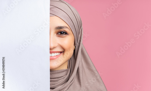 Smiling arabic girl standing behind blank advertisement board, covering half face © Prostock-studio