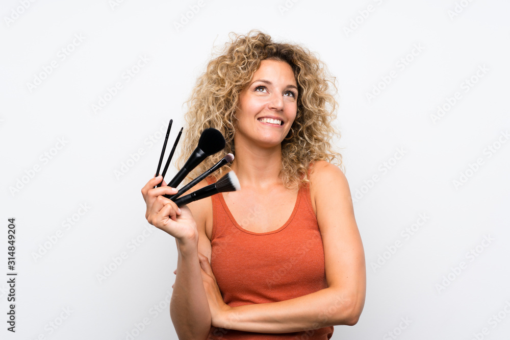 Young blonde woman with curly hair holding a lot of makeup brush looking up while smiling Young blonde woman with curly hair holding a lot of makeup brush looking up while smiling