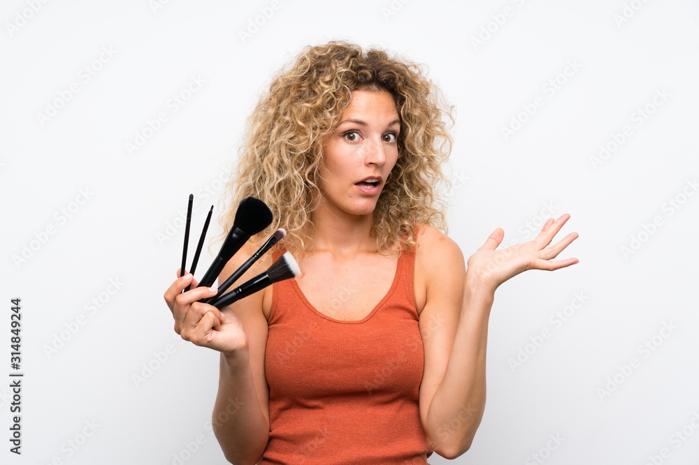 Young blonde woman with curly hair holding a lot of makeup brush with shocked facial expression Young blonde woman with curly hair holding a lot of makeup brush with shocked facial expression