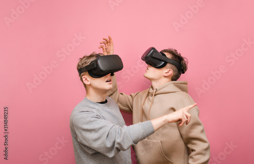 Two surprised guys in VR glasses stand on a pink background looking away and pointing their finger. Two others watching in headset of virtual reality, they are shocked. Copy space