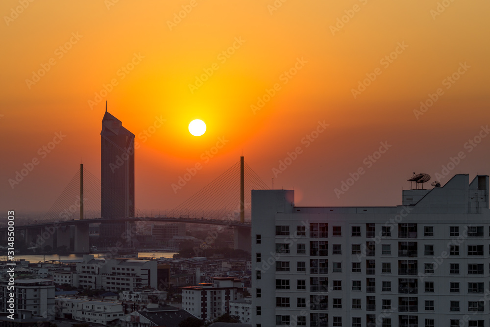 The background of the big sun that is hiding in the evening on the bridge over the river, is naturally beautiful and there are many tall buildings in the area below.