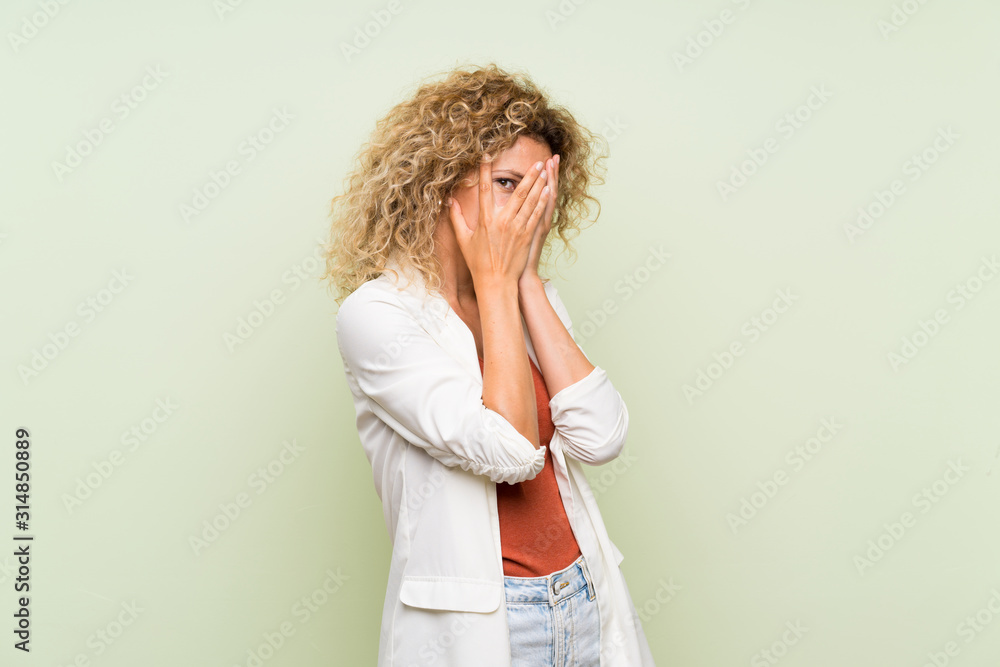 Young blonde woman with curly hair over isolated green background covering eyes and looking through fingers Young blonde woman with curly hair over isolated green background covering eyes and looking through fingers