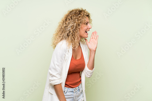 Young blonde woman with curly hair over isolated green background shouting with mouth wide open