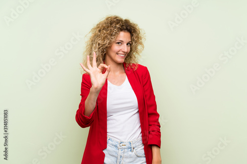 Young blonde woman with curly hair over isolated green background showing ok sign with fingers