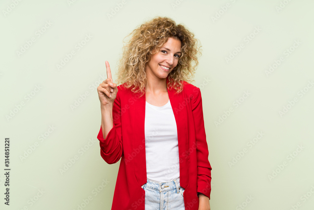 Young blonde woman with curly hair over isolated green background showing and lifting a finger in sign of the best Young blonde woman with curly hair over isolated green background showing and lifting a finger in sign of the best