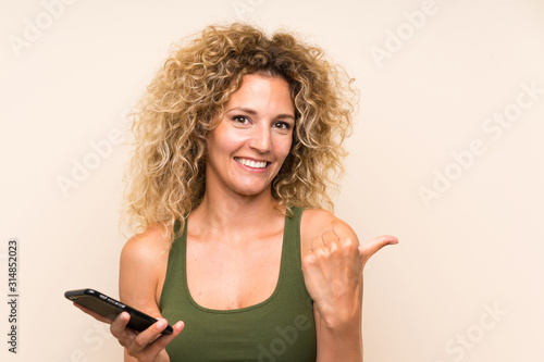 Young blonde woman with curly hair using mobile phone pointing to the side to present a product